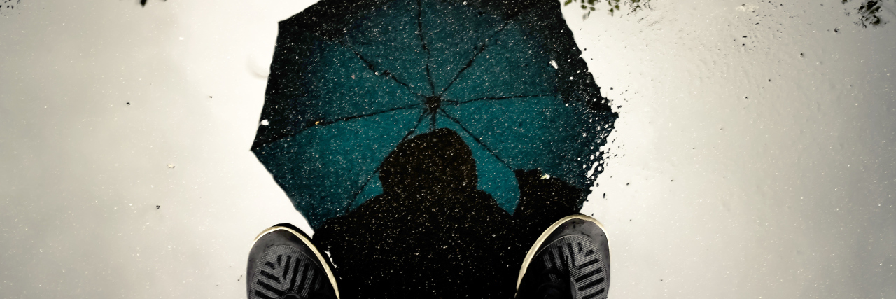 A reflection in a puddle of a person standing with an umbrella open and above their head.