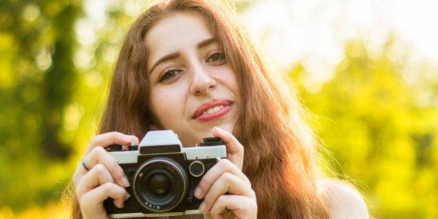 A young woman holding a camera, while she smiles.