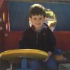 Little boy sitting in an outdoor play train, he is sitting holding a large yellow wooden wheel.
