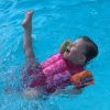 Little girl with a disability swimming, she wears a pink suit that has floating devices and orange swimmingwings and she is kicking a leg up in delight.