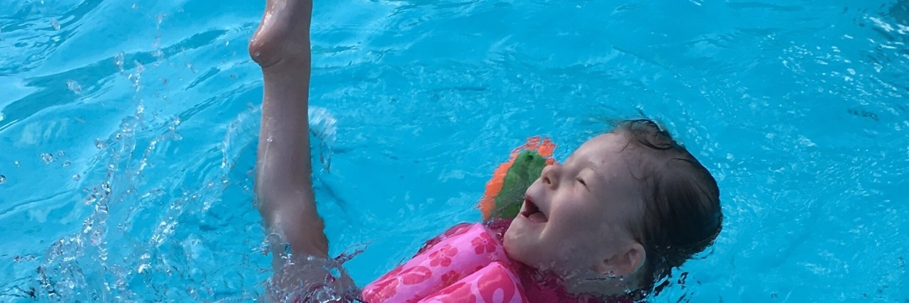 My Child's List of Medical Conditions Should Not Be What Defines Her Little girl with a disability swimming, she wears a pink suit that has floating devices and orange swimmingwings and she is kicking a leg up in delight.