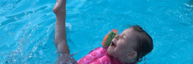 Little girl with a disability swimming, she wears a pink suit that has floating devices and orange swimmingwings and she is kicking a leg up in delight.