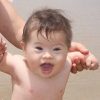 Little boy with Down syndrome at the beach, holding on to sister's hands and wearing a blue swim suit