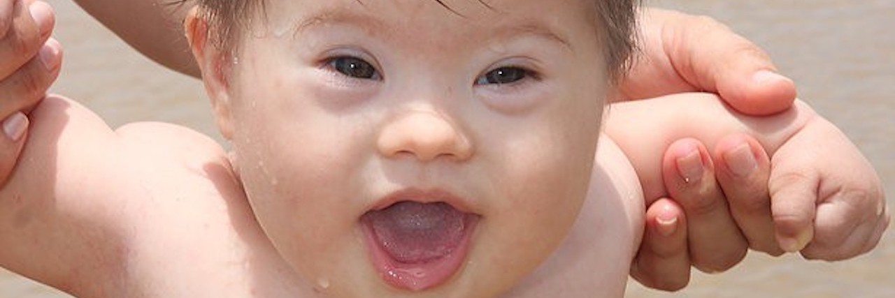 My Brother With Down Syndrome Changed Me Forever Little boy with Down syndrome at the beach, holding on to sister's hands and wearing a blue swim suit