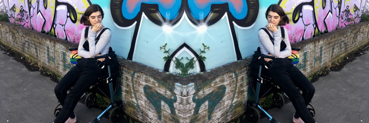 Charlie sitting on her rollator, with her eyes closed. She is in front of a wall with blue, black and purple street art on it. The entire image is mirrored down the middle.