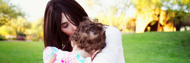 Mother sitting on blanket outdoors hugging and kissing her daughter. Daughter's face is on mother's chest.