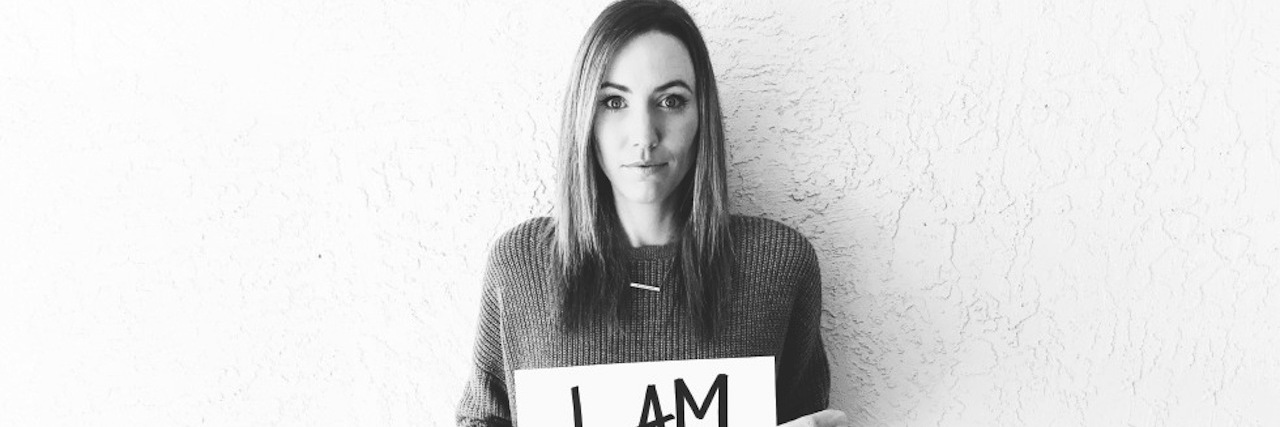 Dear Endometriosis, I Haven't Lost My Voice A black and white image of the writer standing against a white wall, holding a sign that reads, "I am 1 in 10."