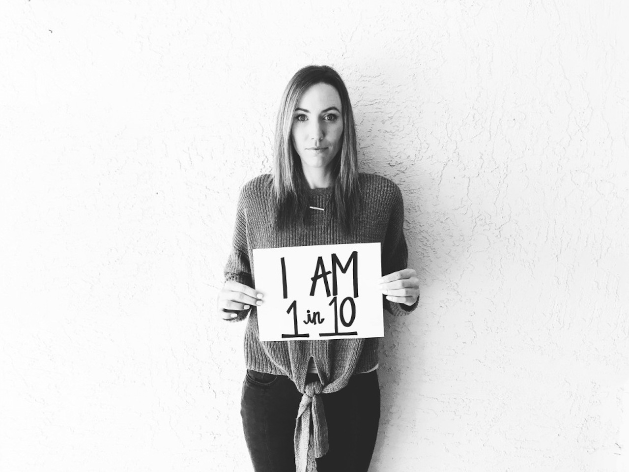 A black and white image of the writer standing against a white wall, holding a sign that reads, "I am 1 in 10."