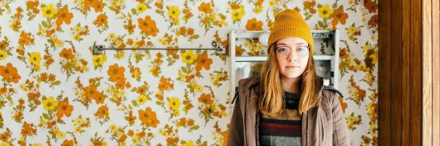woman with hat and glasses stands in bathroom with floral wallpaper