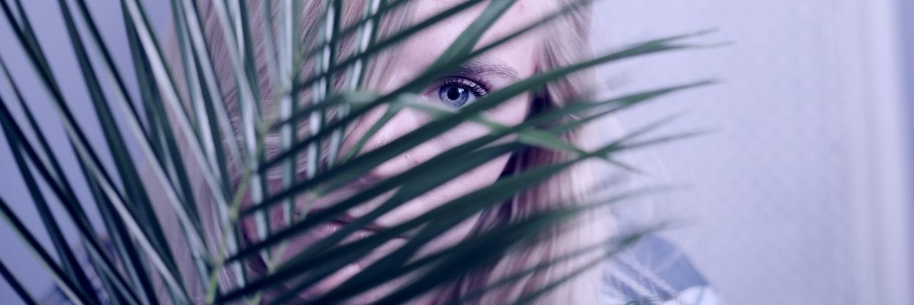 young girl with blonde hair and blue eyes hiding behind plant