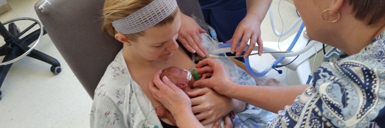 Mother holding preemie baby for first time, two nurses' hands visible as they help with the monitors, wires, and support for baby