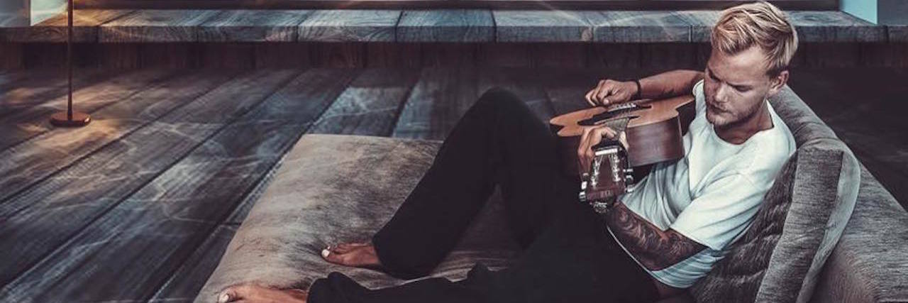 How I Apply Avicii's Song 'Trouble' to My Life With Chronic Illness A picture of Avicii playing his guitar on a bed, with a window showing ocean life like a whale – from the perspective of being underwater.