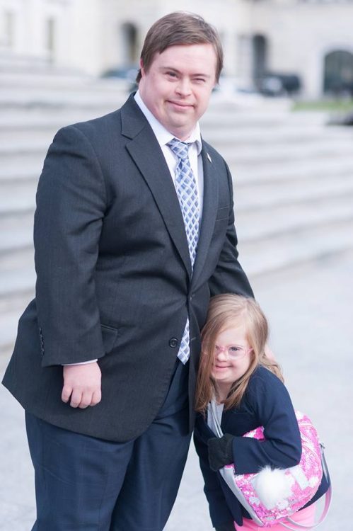 Adrian posing with little girl with Down syndrome hugging side by side. Adrian wears a suit and stands before a government building