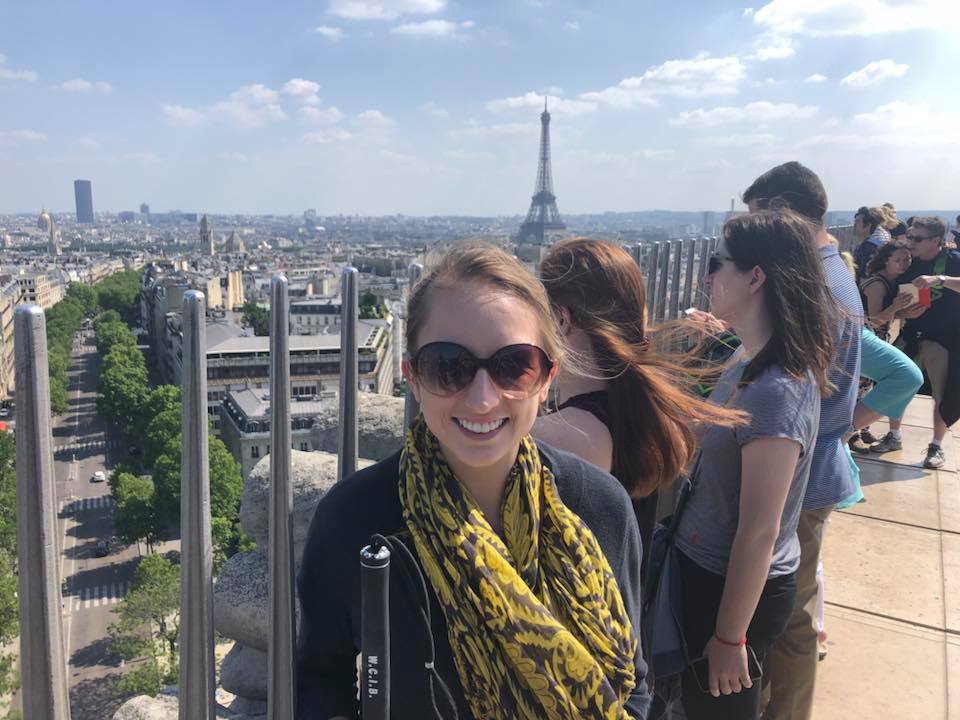 Shannon Reyenga stands on top of an observation deck, in the distance behind her is the Eiffel Tower. She is wearing sunglasses and a yellow scarf. She is holding a cane. 