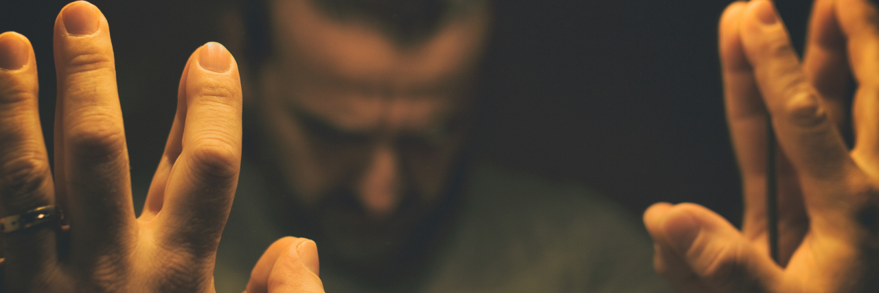 When the 5 Stages of Grief Are Caused by Illness A man in a dimly lit room, his hands up to a mirror, showing the reflection of his face looking down.