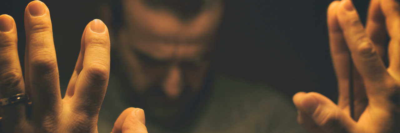 A man in a dimly lit room, his hands up to a mirror, showing the reflection of his face looking down.