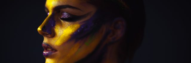 A picture of a woman with paint on her face and body, mostly yellows and some purple.
