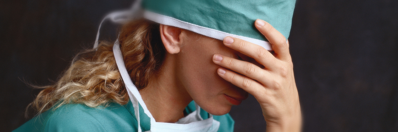 Why Getting Sick While Working in Healthcare Doesn't Make Me 'Lucky' A female medical professional wearing scrubs, with her head in her hand, in front of a dark background.