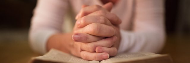 Unrecognizable woman praying with hands clasped together on her Bible. Close up.