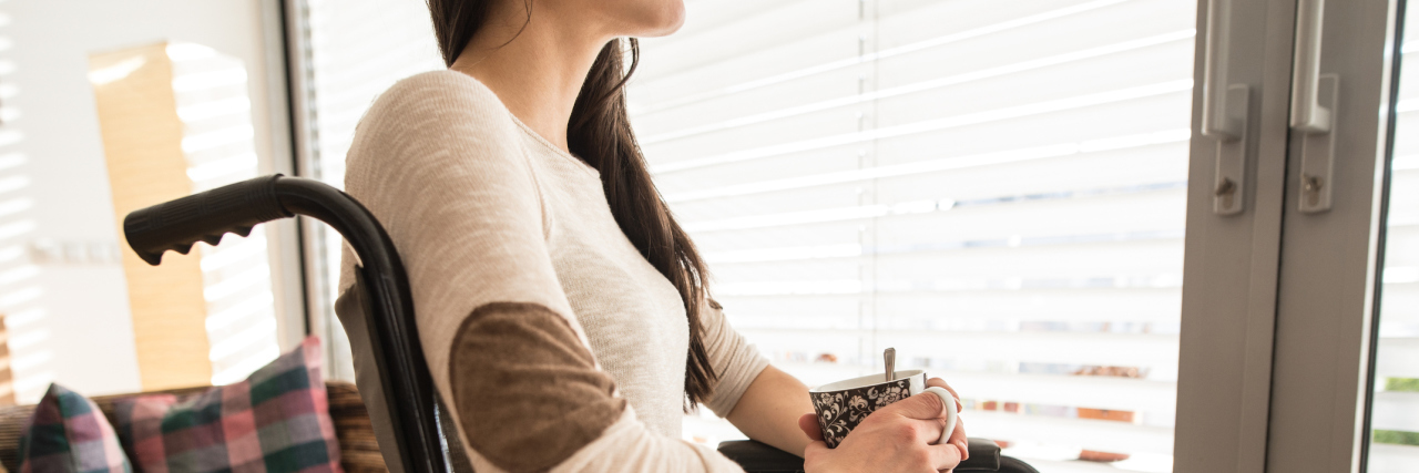The Anxiety That Comes With Using a Wheelchair for the First Time A woman sitting in a wheelchair, holding a cup of tea, while looking out the window.