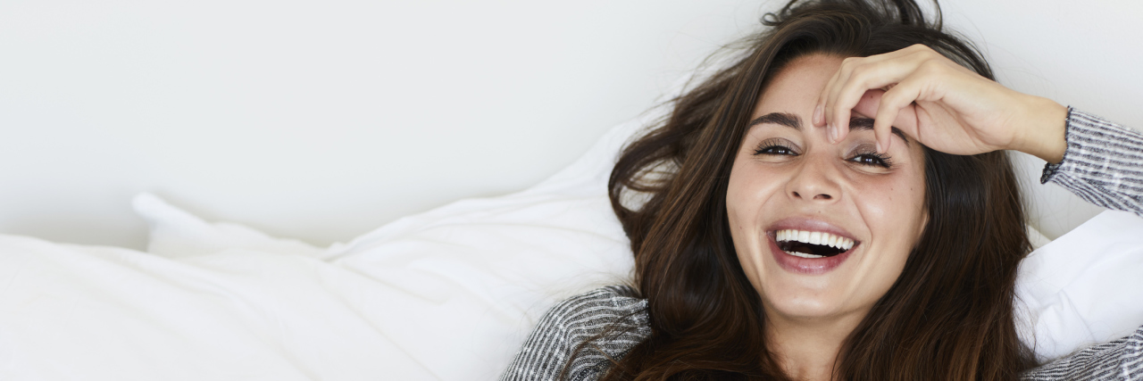 How I'm Finding Humor in My Chronic Illnesses A woman laying on a bed with white sheets, holding a cup of coffee, while laughing.