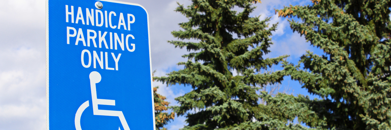 A picture of a handicap parking sign with trees and a blue sky in the background.