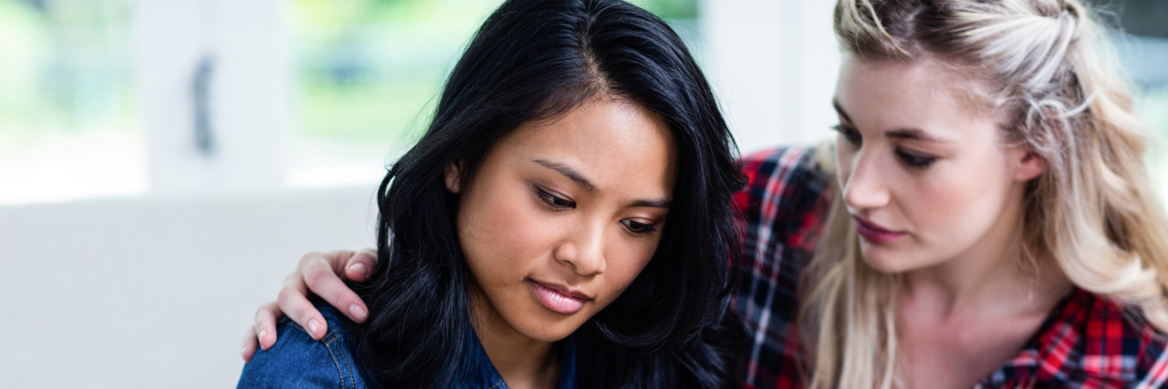 A photo of two friends, one with her arm around the other while they talk.