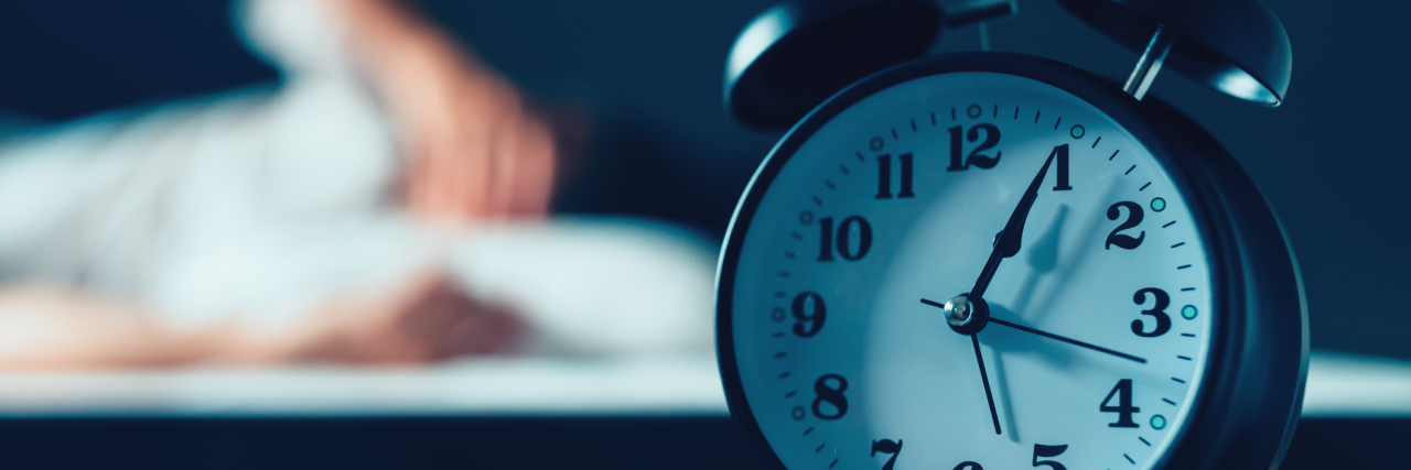 Why the Saying 'Everyone Has 24 Hours in a Day' Is Ableist alarm clock on the nightstand with a person in the background struggling to sleep