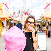 A woman eating pink cotton candy while outside at an amusement park.