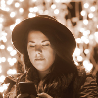 A woman looking at her phone outside, with beautiful lights hanging behind her.