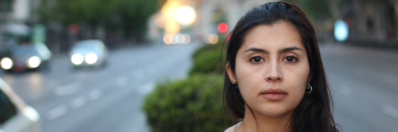 The Difference Between Accepting and Coming to Terms With Your Illness A picture of a Latina woman with a serious facial expression, standing outside in a median.