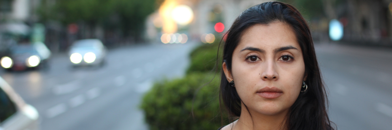 A picture of a Latina woman with a serious facial expression, standing outside in a median.