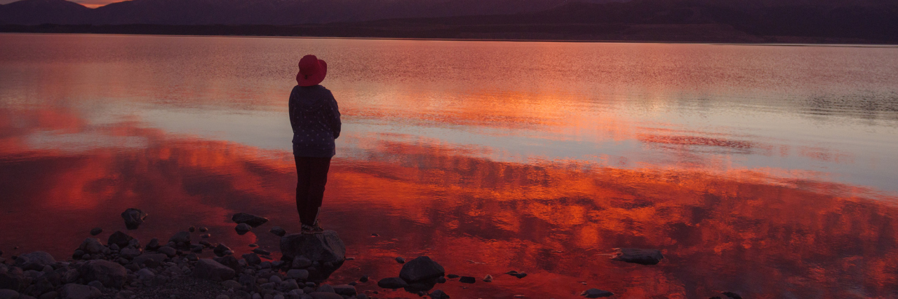 A picture of a woman watching a sunset, which is reflecting on a body of water in front of her.