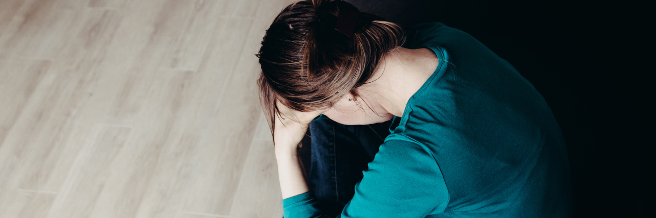 What I Want People to Understand About Myalgic Encephalomyelitis A picture of a woman sitting on her couch, with her head facing down and resting in her hands.