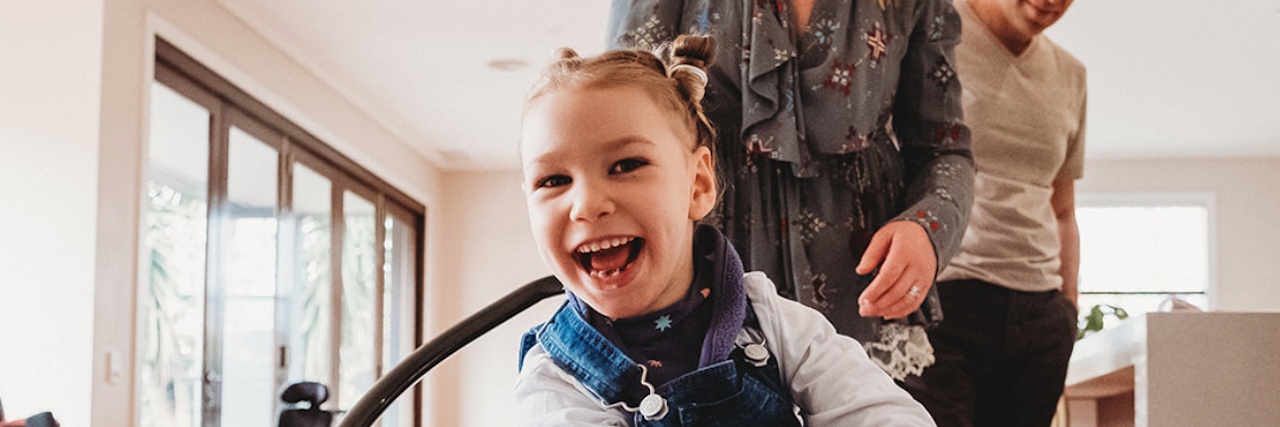 The Powerful Community You Connect With When Your Kid Has a Disability Child using a walker smiling coming towards camera, mom and dad blurry in the background