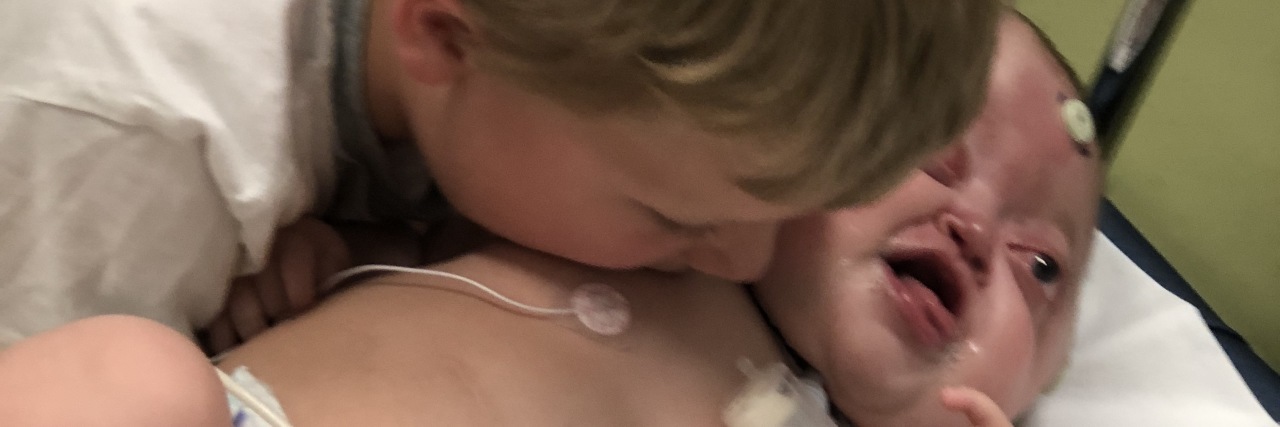 Please Don’t Pray for My Daughter With a Disability to Be Healed Little baby with disabilities in hospital bed, brother is leaning over and kissing her