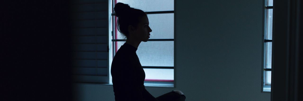 Shadow outline of a woman sitting on her bed. There is pale light coming through the window behind her. Photo by Ben Blennerhassett on Unsplash