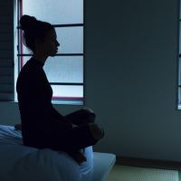 Shadow outline of a woman sitting on her bed. There is pale light coming through the window behind her. Photo by Ben Blennerhassett on Unsplash