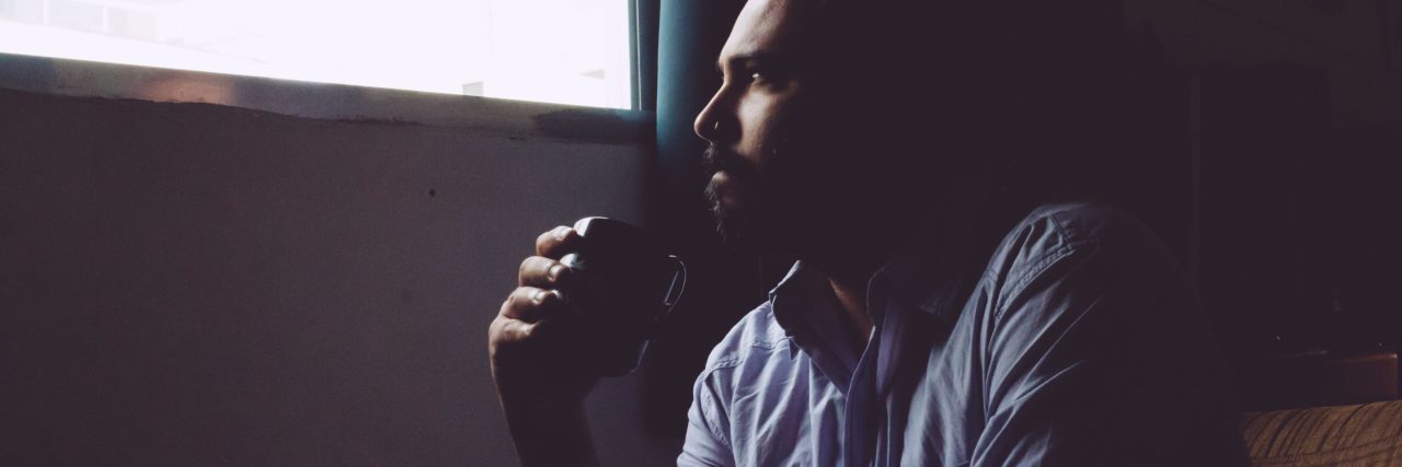 man in dark room drinking coffee and looking out window