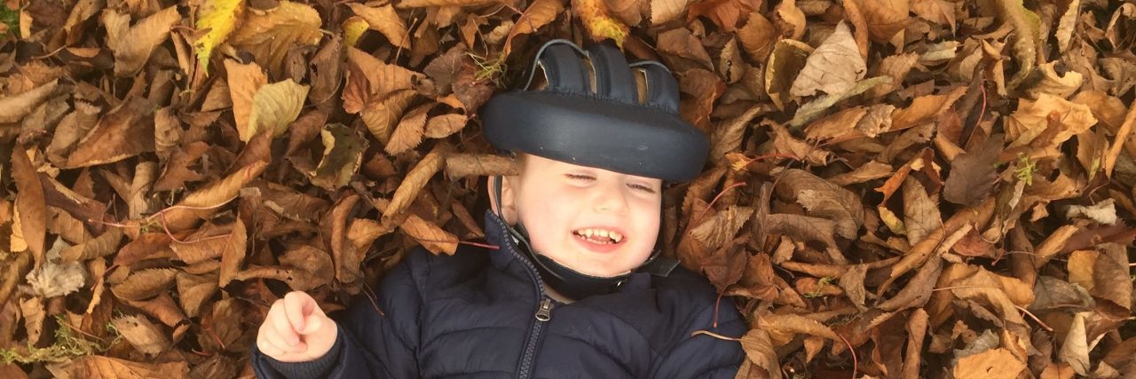 How My Son's Medical Condition Impacted My Parenting Style Little boy wearing a medical helmet smiling and laying on a pile of leaves