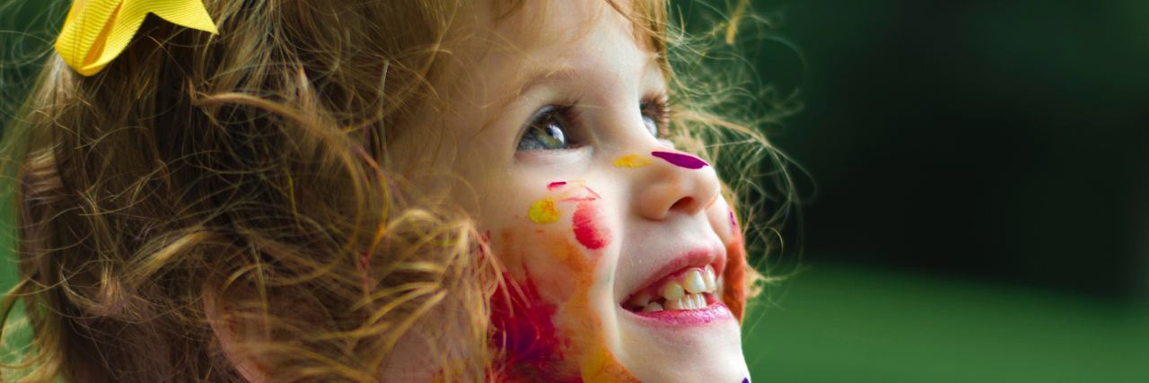 Little girl covered in colorful pain all over face, nech, white clothes. She is smiling off to the side
