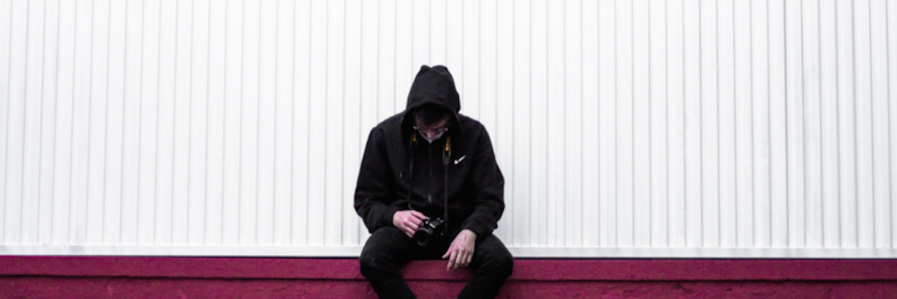 A man in a black hoodie and jeans sits on a red wall, hood pulled up, looking lethargically down at the camera he's holding. 15 Behaviors We Don't Always Recognize Are Self-Harm