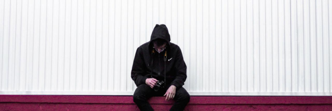 15 Behaviors We Don't Always Recognize Are Self-Harm A man in a black hoodie and jeans sits on a red wall, hood pulled up, looking lethargically down at the camera he's holding. 15 Behaviors We Don't Always Recognize Are Self-Harm