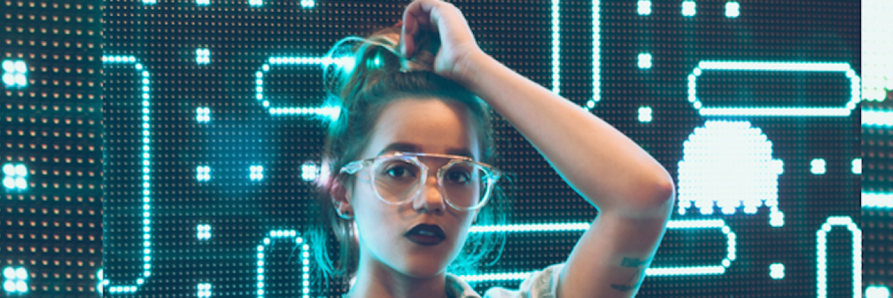 A woman with pink-rimmed glasses stands in front of a large 8-bit Pacman game. 17 Things People With Borderline Personality Disorder Do That Are Code for 'Don't Leave Me'