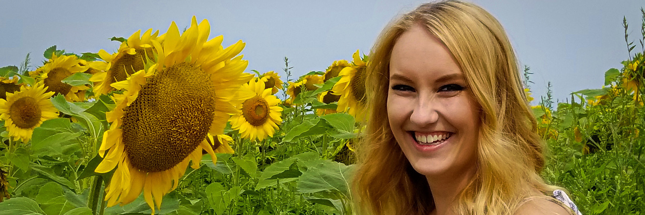 Please Stop Holding It Against Me That I Can Mask My Pain A picture of the writer standing next to a sunflower in a field.