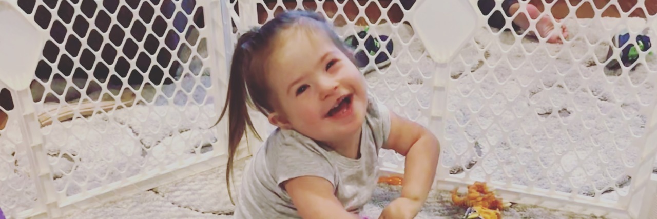 As the Mom of a Child With a Disability, Please Don't Say This Phrase Little girl with Down syndrome sitting on floor, smiling at camera with baby gate behind her