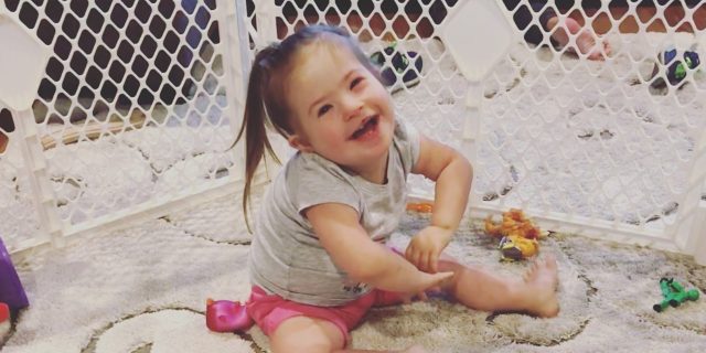 Little girl with Down syndrome sitting on floor, smiling at camera with baby gate behind her