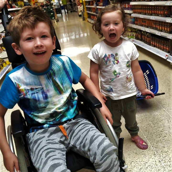 Picture of two kids, boy in a wheelchair and little sister next to him walking and opening her mouth wide