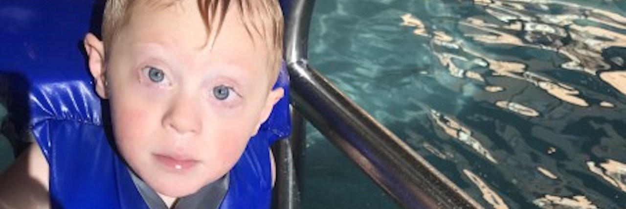 Why I Don't Always Think About My Son Having Down Syndrome Boy with Down syndrome looking at camera, he is in a pool wearing a blue life vest