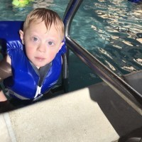Boy with Down syndrome looking at camera, he is in a pool wearing a blue life vest
